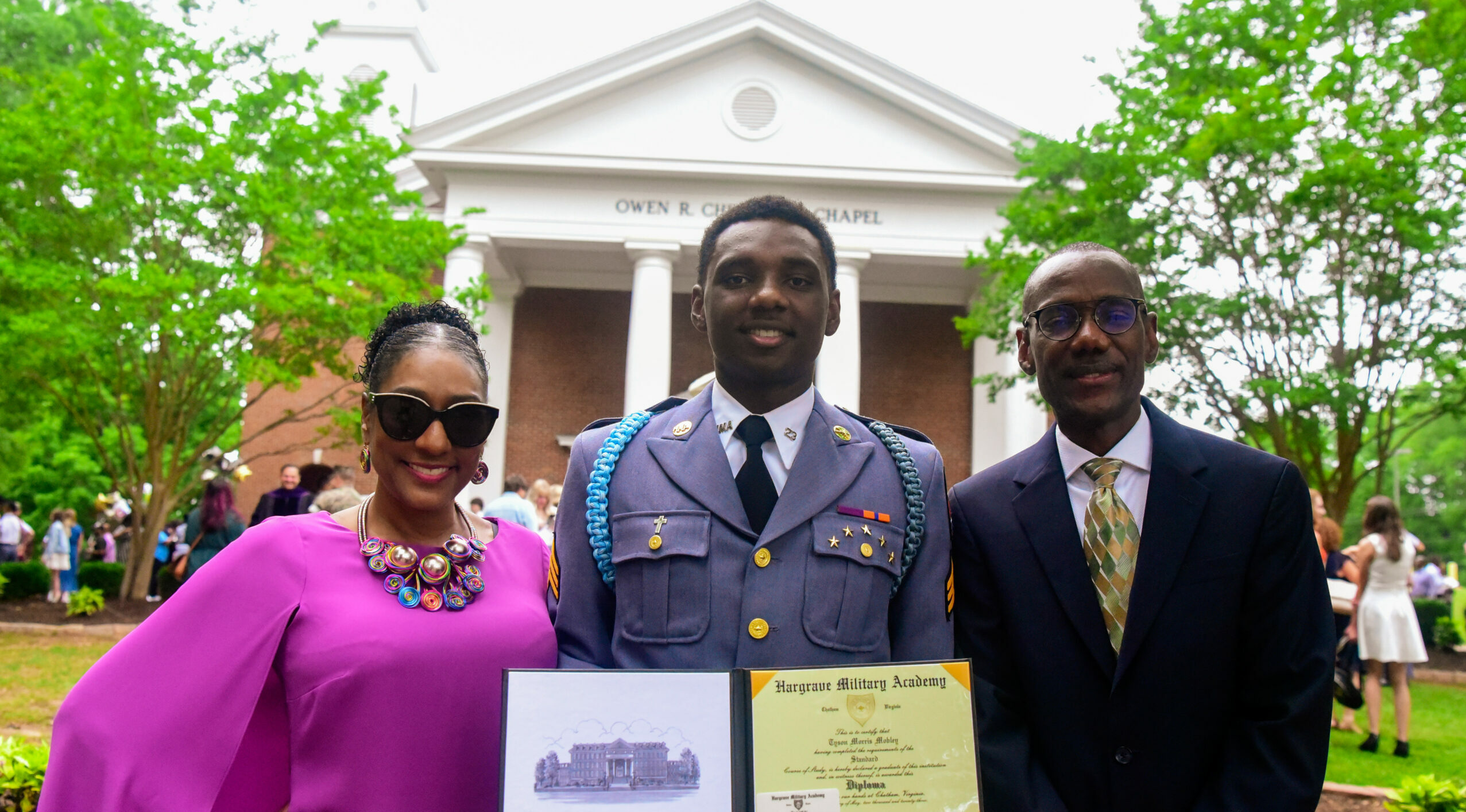 Cadet with his parents smiling at the viewer in front of the chapel after graduation.