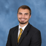 May show: A man in a dark suit, white shirt, and striped yellow tie smiles at the camera against a blue studio background.