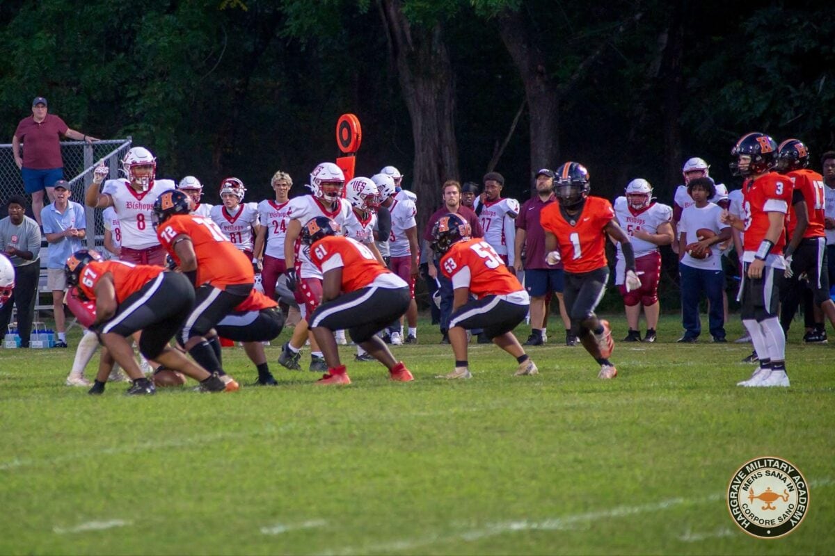 May show: Two high school football teams face off on a grassy field at night, with one team in red and black uniforms and the other in white. Coaches and players watch from the sideline.