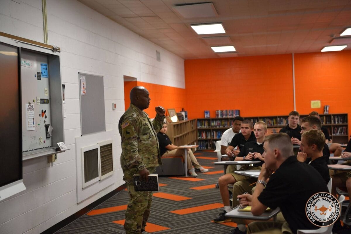 May show: A uniformed military instructor stands at the front of a classroom speaking to students seated at desks, with bookshelves and a whiteboard in the background.