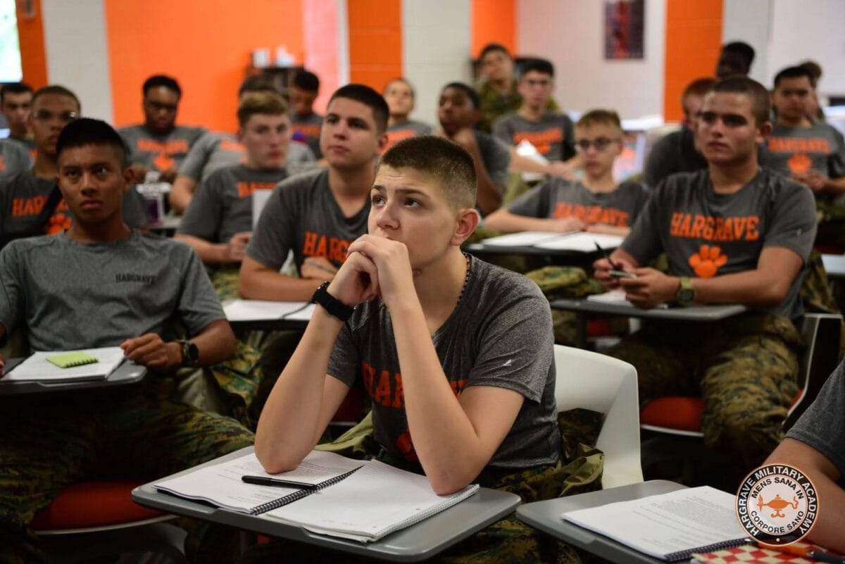 May show: A group of students in matching uniforms sit at desks in a classroom, attentively listening and taking notes.